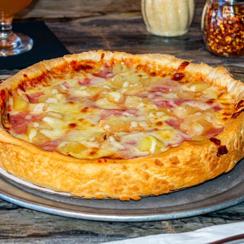 A close-up of a Sicilia's Pizzeria deep-dish pizza with ham and pineapple on a metal tray.